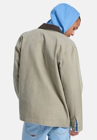 Ανοιχτό πράσινο βαμβακερό μπουφάν με καφέ κοτλέ γιακά, σε άνετη εφαρμογή με μανσέτες με κουμπιά, φορεμένο πάνω από μια μπλε φούτερ με κουκούλα.