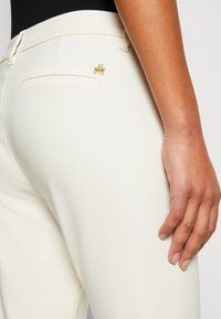 Gros plan d'une personne portant un pantalon blanc ajusté avec une épingle logo "M" dorée au-dessus de la poche arrière et un haut noir.