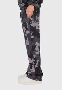 Osoba ubrana w szare i czarne spodnie w kamuflażu cyfrowym oraz białe sneakersy, stojąca bokiem z ręką opartą na udzie.