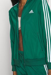 Kobieta w zielonej kurtce dresowej Adidas i spodniach z białymi paskami, częściowo rozpięta, odsłaniająca biały krótki top pod spodem.