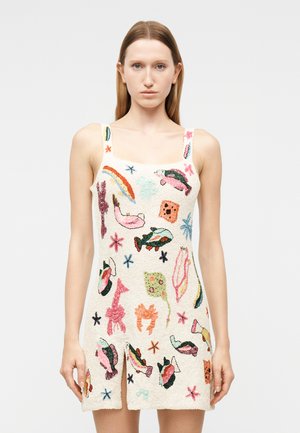 Γυναίκα που φοράει αμάνικο, κρεμ φόρεμα με πολύχρωμες κεντημένες θαλασσινές φιγούρες και λουλουδάτα σχέδια, στέκεται μπροστά σε απλό φόντο.