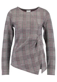 Haut à manches longues gris avec un motif à carreaux comprenant des lignes noires, blanches et rouges. Ourlet asymétrique et détails en tissu texturé.