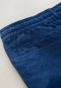 Gros plan sur la ceinture d'un pantalon en tissu bleu avec un élastique et une poche passepoilée simple sur un fond clair.
