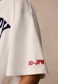 Balta kokvilnas T-kreklu ar tumši zilu uzdrukātu tekstu un sarkanu izšūtu "SD-JPN" uz piedurknes. Īsām piedurknēm ar brīvu piegriezumu.