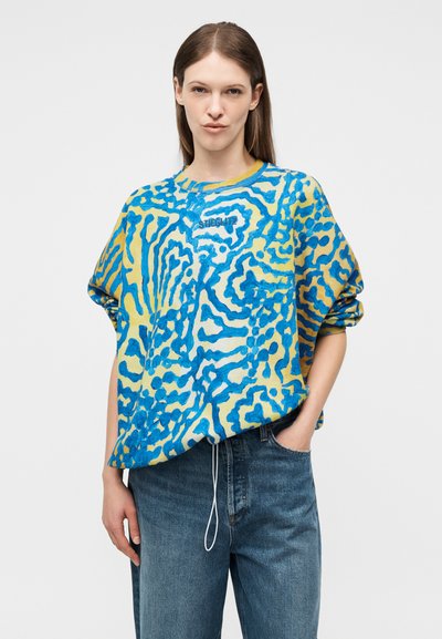 Γυναίκα που φοράει πουλόβερ με μπλε και κίτρινο αφηρημένο μοτίβο και την επιγραφή "STIEGLITZ", συνδυασμένο με άνετο μπλε τζιν, ποζάροντας με το ένα χέρι στην τσέπη.