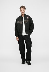 Tânăr îmbrăcat cu o jachetă de curse neagră cu inscripția "Hilfiger Racing Club" și pantaloni cargo, cămașă albă și pantofi negri, pe un fundal alb.