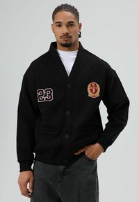 Zwarte cardigan gemaakt van zachte stof, met een knoopsluiting, twee voorzakken, geborduurde accenten en het voetbalnummer "23" op de mouw.