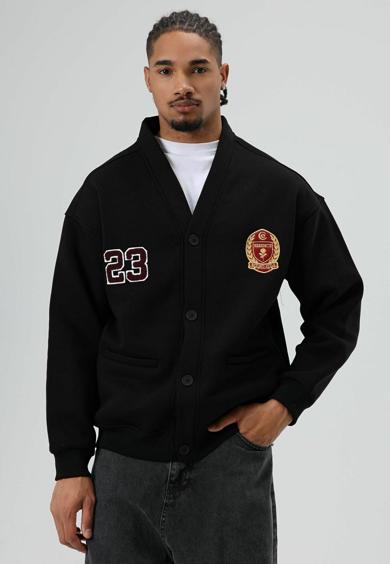 Zwarte cardigan gemaakt van zachte stof, met een knoopsluiting, twee voorzakken, geborduurde accenten en het voetbalnummer "23" op de mouw.