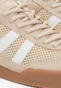 Beige sportschoen met een bovenwerk van suede en mesh, witte accentstrepen, platte beige veters en een gumrubberen buitenzool. Close-up weergave.