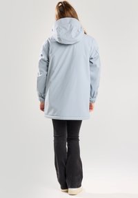 Vaaleansininen vedenpitävä takki huppulla, elastisilla hihansuilla ja hieman pidennetty takaa, käytetty mustien levyhaalarien ja valkoisten kenkien päällä.