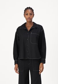 Svart button-up skjorte med krage, med hvite kontrastsømmer, lange ermer og en brystlomme. Glatt stofftekstur.