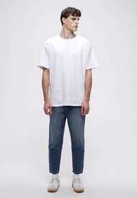 Hvit bomulls-T-skjorte med løs passform, kombinert med blå slankende denimjeans. Skoene er hvite med brun såle, plassert mot en nøytral bakgrunn.