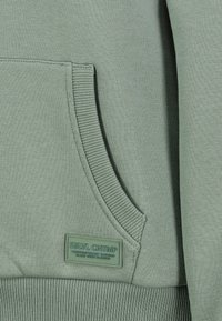 Grüner Baumwollpullover mit Kängurutasche, gerippten Bündchen und Saum sowie einem strukturierten Etikett mit eingeprägtem Text. Weicher Stoff.