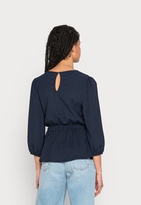 Top peplum azul marinho com decote redondo, mangas bufantes e detalhe de abertura nas costas, feito de tecido texturizado. Combinado com jeans de lavagem clara.