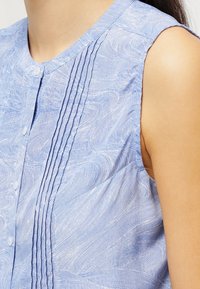 Blusa senza maniche di un blu chiaro con un motivo a onde, caratterizzata da pieghe verticali e un collo rotondo con chiusure a bottoni, realizzata in un tessuto strutturato.