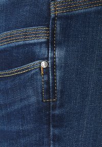 Nära bild av blå denimtyg med gula sömmar och en liten metallnitar i hörnet på en jeansficka.