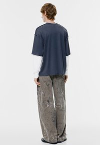 Túlméretezett sötétkék póló egy fehér hosszú ujjú ing fölött, világosszürke, mintás, bőszárú nadrággal párosítva, látható varrással és zsebekkel.