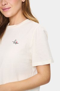 Kreminė trumpa rankovė marškinėlių iš minkštos medžiagos, ant kairiojo krūtinės regiono yra mažas juodas ir raudonas išsiuvinėtas "meilės" dizainas.