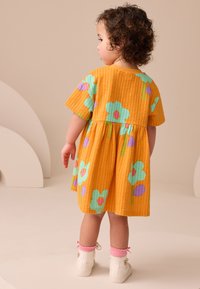 Vestido amarillo de algodón con mangas cortas, con grandes flores turquesas y moradas, tela texturizada y cintura fruncida. Zapatos blancos con calcetines rosas.