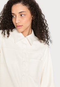 Camisa de couro sintético branca cremosa com gola, fecho de botões, bolso frontal e detalhes de costura visíveis.