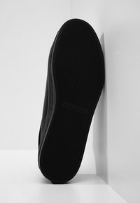 Baskets noires avec une semelle en caoutchouc, présentant un design texturé et un logo discret sur le côté. La semelle est inclinée sur un fond blanc.