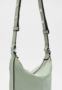 Grünlederhandtasche mit einzigartiger asymmetrischer Form, mit schwarzer Kante, verstellbarem Tragegurt und metallischen Hardware-Akzenten.