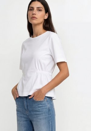 Žena s dlhými tmavými vlasmi, oblečená v biele krátky rukáv s naťahovaným pásom a modré džínsy, stojaca s rukami v vreckách na svetlom pozadí.