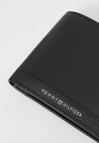Portefeuille en cuir noir avec une texture lisse et des coutures horizontales. Comprend le logo "TOMMY HILFIGER" en blanc sur le devant.