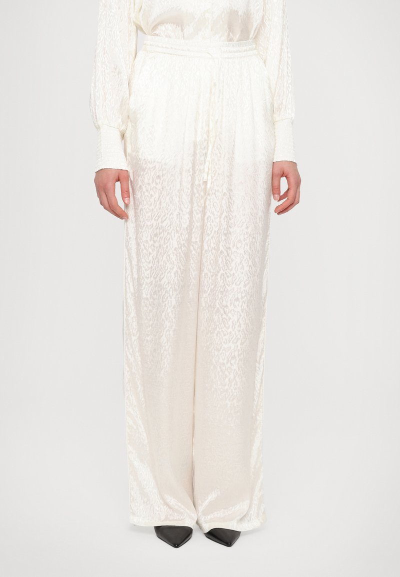 White, flowy pants made of textured fabric with a subtle pattern. Features a drawstring waist and long, wide legs. Worn with black pointed shoes.