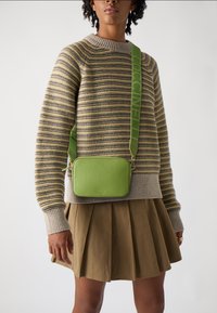 Zelená texturovaná kožená crossbody taška s konturovaným tvarem, zdobená logovými popruhy a zipovým uzávěrem, nošená přes pruhovaný svetr a plisovanou sukni.