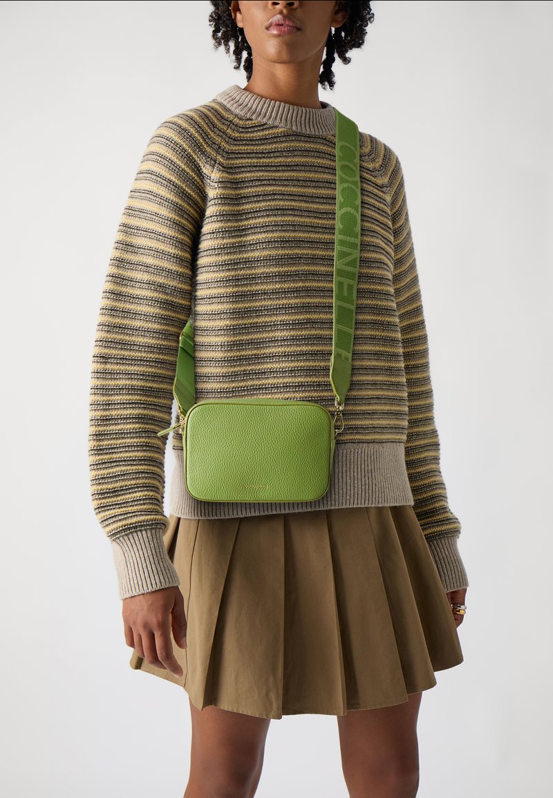 Zelená texturovaná kožená crossbody taška s konturovaným tvarem, zdobená logovými popruhy a zipovým uzávěrem, nošená přes pruhovaný svetr a plisovanou sukni.