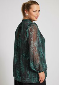 Blusa trasparente verde con un motivo a stampa leopardata in tonalità scure e chiare. Presenta maniche a sbuffo e un colletto alto.
