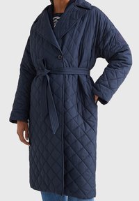 Gepolsterter dunkelblauer Mantel mit tailliertem Gürtel, breitem Kragen und großem Aufnäher am Ärmel. Verfügt über diagonale Nähte und zwei Seitentaschen.