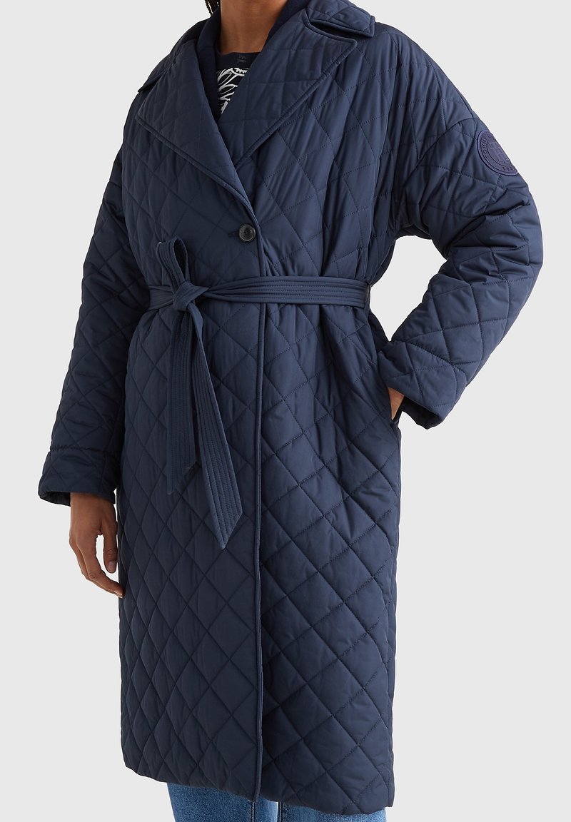 Gepolsterter dunkelblauer Mantel mit tailliertem Gürtel, breitem Kragen und großem Aufnäher am Ärmel. Verfügt über diagonale Nähte und zwei Seitentaschen.