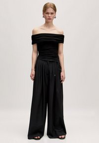 Zwarte off-shoulder top met geplooide textuur en een strikdetail bij de taille, gecombineerd met zwarte broek met wijdlopende pijpen en platte sandalen.
