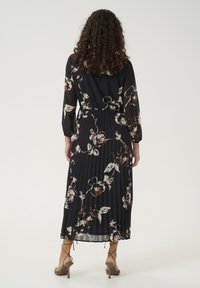 Melna puķaina maksi kleita ar garām piedurknēm, pleatēta tekstūra un brūnas puķu rakstainas. Nēsāta ar siksniņu zaļiem papēžiem, redzams aizmugures skats.