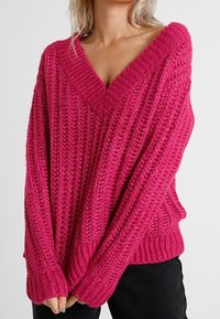 Pull en tricot fuchsia avec un col en V profond, présentant une texture côtelée épaisse, des épaules tombantes et une coupe oversize. Comprend deux poches latérales.