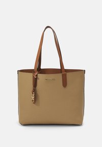 Béžová taška s texturou a hnědými koženými popruhy, která má pozlacený logo akcent a malou ozdobu připevněnou k popruhu.