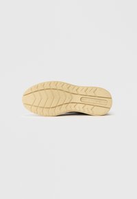 Gummi-Außensohle mit einem Zickzackprofil in Hellbeige, ausgestattet mit geprägtem Branding und einer glatten, flachen Oberfläche.
