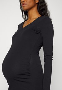 T-shirt de maternidade preto de mangas longas com decote em V, detalhes franzidos laterais e tecido macio e elástico projetado para acomodar uma barriga em crescimento.