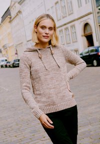 Beige kötött pulóver kapucnival, fából készült gombokkal, bordázott mandzsettával és finom textúrájú mintával. Sötét nadrággal viselve.