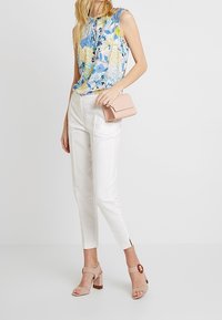 Haut sans manches floral en bleu et jaune, pantalon cropped blanc, pochette rose et sandales à talons bloc beiges avec un accent circulaire.