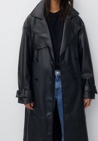 Femme portant un long trench en cuir noir, une chemise noire et un jean bleu, se tenant devant un fond clair.