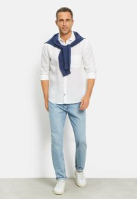 Weißes Leinenhemd mit langen Ärmeln und einer Brusttasche, kombiniert mit hellblauen Denim-Jeans und weißen Sneakers. Dunkelblauer Pullover um den Hals drapiert.