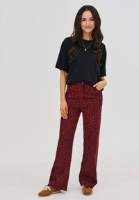 Čierne tričko s krátkym rukávom skombinované s červenými nohavicami s leopardím vzorom a vysokým pásom; model stojí s rukami v vreckách a má na sebe hnedé topánky.