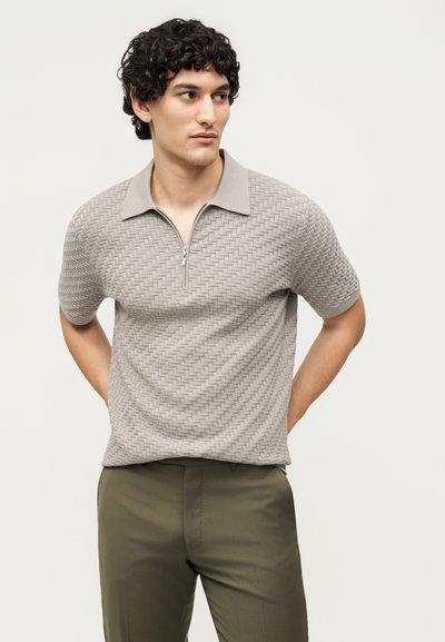 Άνδρας με σγουρά μαλλιά που φοράει υφασμάτινη ανοιχτογκρι κοντομάνικη πόλο με φερμουάρ και χακί παντελόνι, κοιτάζει προς τα πλάγια με τα χέρια πίσω από την πλάτη.