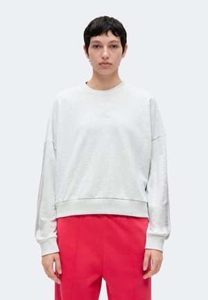 Person med kort mørkt hår iført en lysegrå langærmet sweatshirt og klare røde bukser står foran en ensfarvet baggrund.