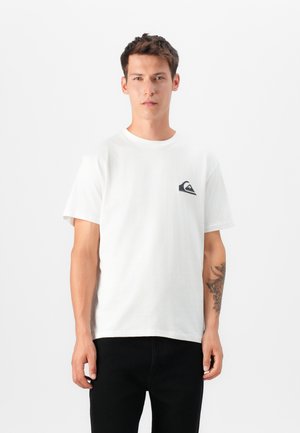 Hvid bomulds T-shirt med rund hals, korte ærmer og et lille sort logo-patch på venstre bryst. Båret med mørke jeans.