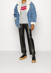 Džínová bunda s umělým kožešinovým límcem, šedé tričko Levi's, černé kožené kalhoty a hnědé platformové boty. Neformální streetwearové oblečení.