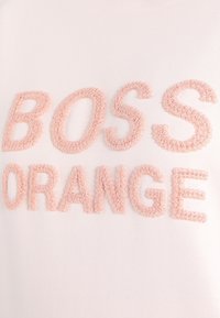 Texto bordado en rosa que dice "BOSS ORANGE" sobre un fondo de tela de color claro, con una textura suave y un diseño de letras en relieve.
